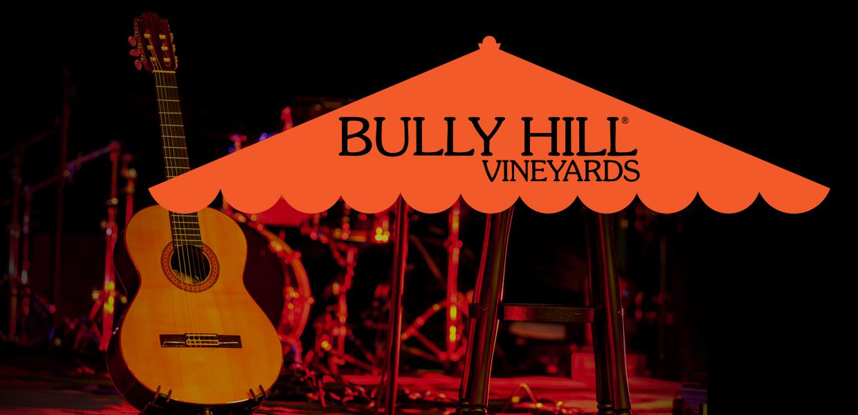 Acoustic guitar next to a stool on a dimly lit stage with "Bully Hill Vineyards" logo overlayed on top.