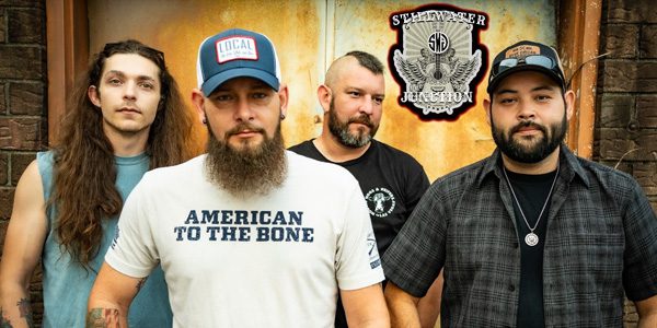 Four men stand in front of a weathered door with a Steel River Junction sign; one wears a cap and a shirt reading "American to the Bone.